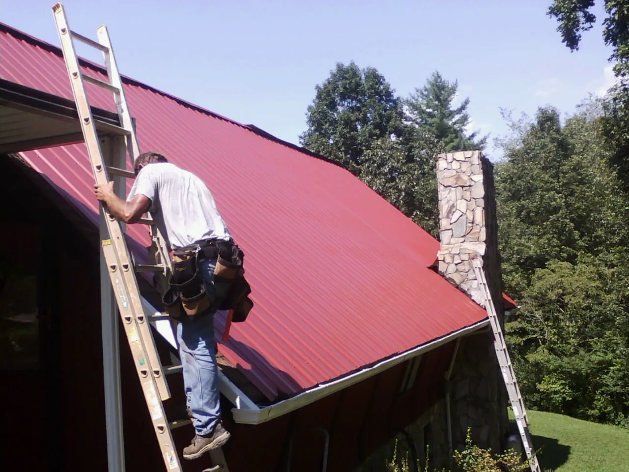 Roofer on a ladder beside a red metal roof and stone chimney — Soffit & Fascia Repair by Roofing Brothers in Verden